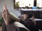 man relaxing on sofa in living room with feet up and reading on a handheld device 