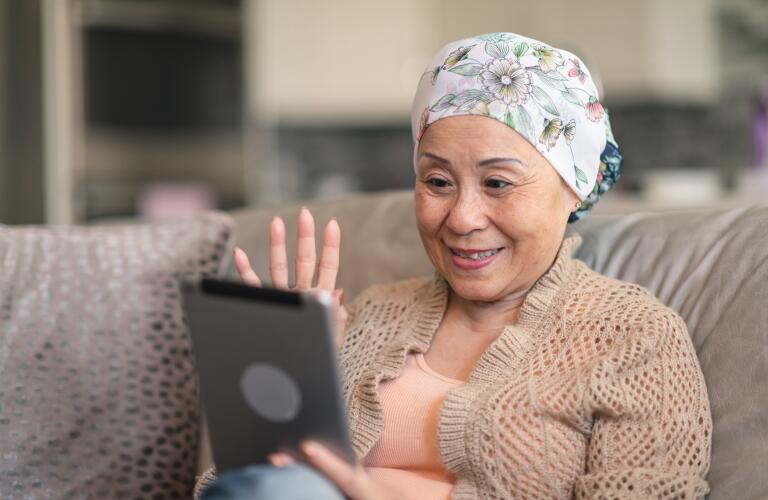 senior Asian female cancer patient waving at tablet and sitting on couch during telehealth appointment