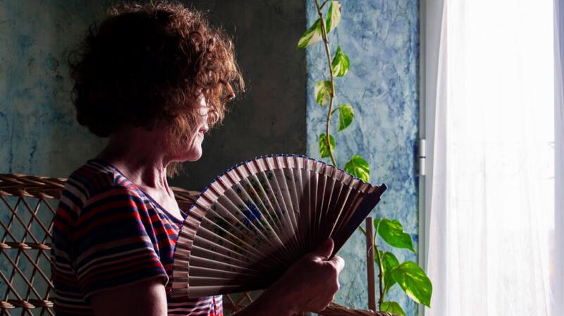 Female standing at a window, fanning herself with a paper fan