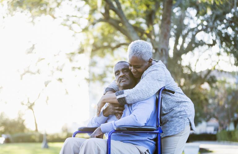 Senior African American man in wheelchair being hugged by wife outdoors in park
