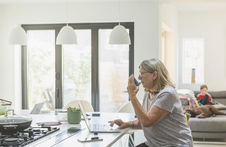 woman sitting at kitchen table in front of laptop using inhaler