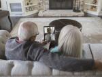 senior Caucasian couple sitting on living room couch holding up tablet with telehealth doctor's appointment