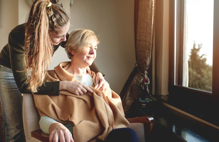 Senior Caucasian woman in chair with young Caucasian female caregiver wrapping her in blanket