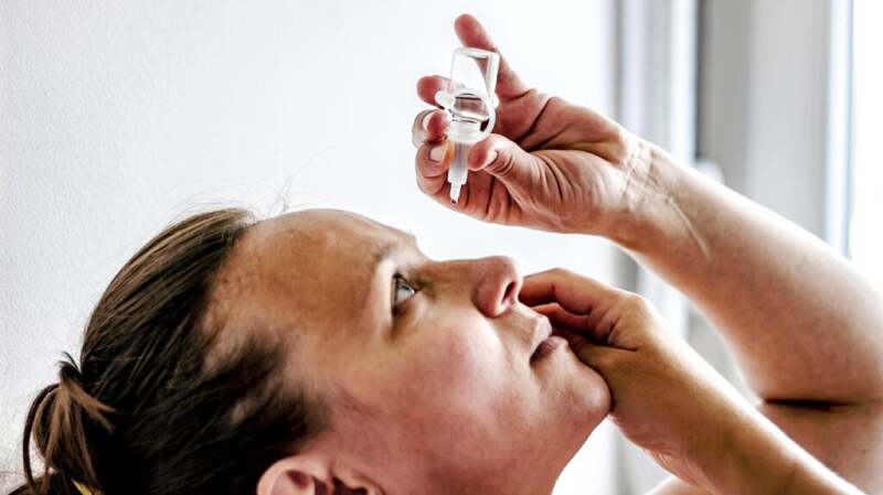 A woman tilts her head back and applies eye drops.