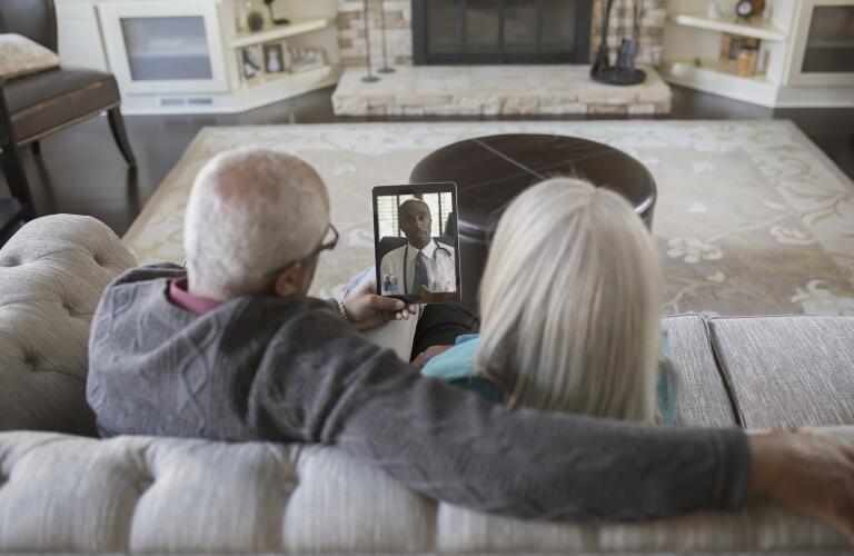 senior Caucasian couple sitting on living room couch holding up tablet with telehealth doctor's appointment