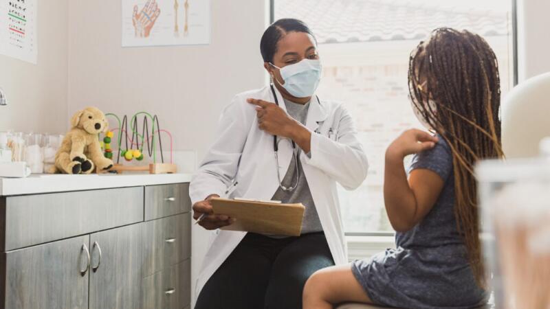 A young child talks to a doctor in a clinic, wearing face masks.