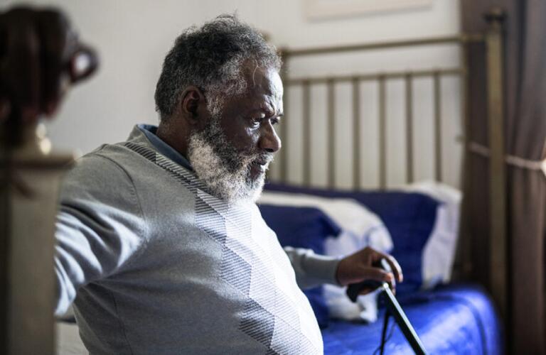 An older man sits on the edge of a bed and uses a cane to support himself.
