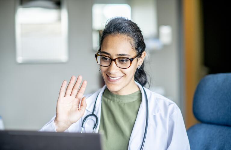 portrait of female doctor waving at computer screen during telehealth appointment