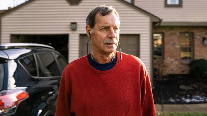 An older male stands in front of a driveway wearing in-ear headphones.