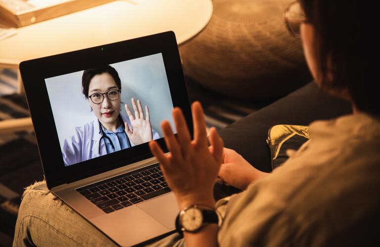 female patient sitting on couch waving during telehealth session with female doctor