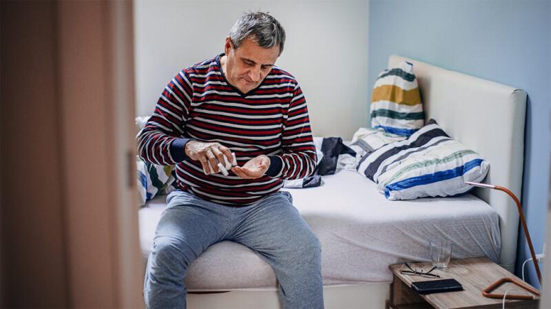 Older man, dressed and sitting on side of bed taking pills