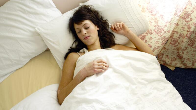 A person lies in bed under a duvet with their eyes closed.