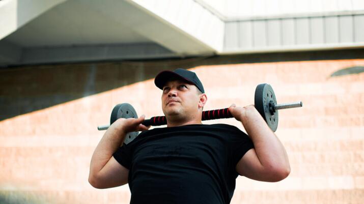 A man holds a barbell with small weight plates over his shoulders, exercising outside in the sun.