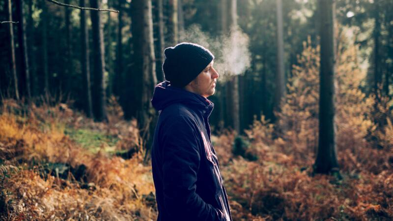 Male stands in a wood wearing a wool hat and coat. His breath vaporizes visibly in the air.