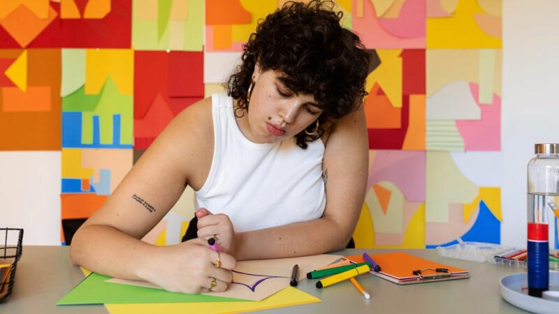 A woman sitting at a table and drawing with her head tilted to the side