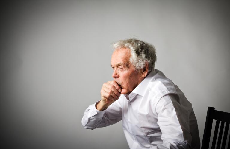 senior male Caucasian sitting in chair coughing against plain gray background