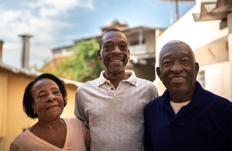 African American parents and adult son stand together outdoors smiling with blue sky in background