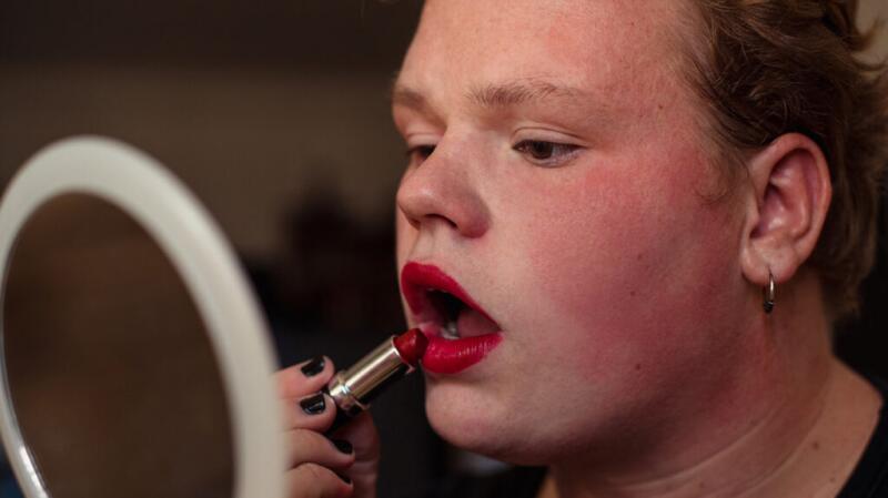 A person with flushed facial skin applies red lipstick while looking in the mirror.